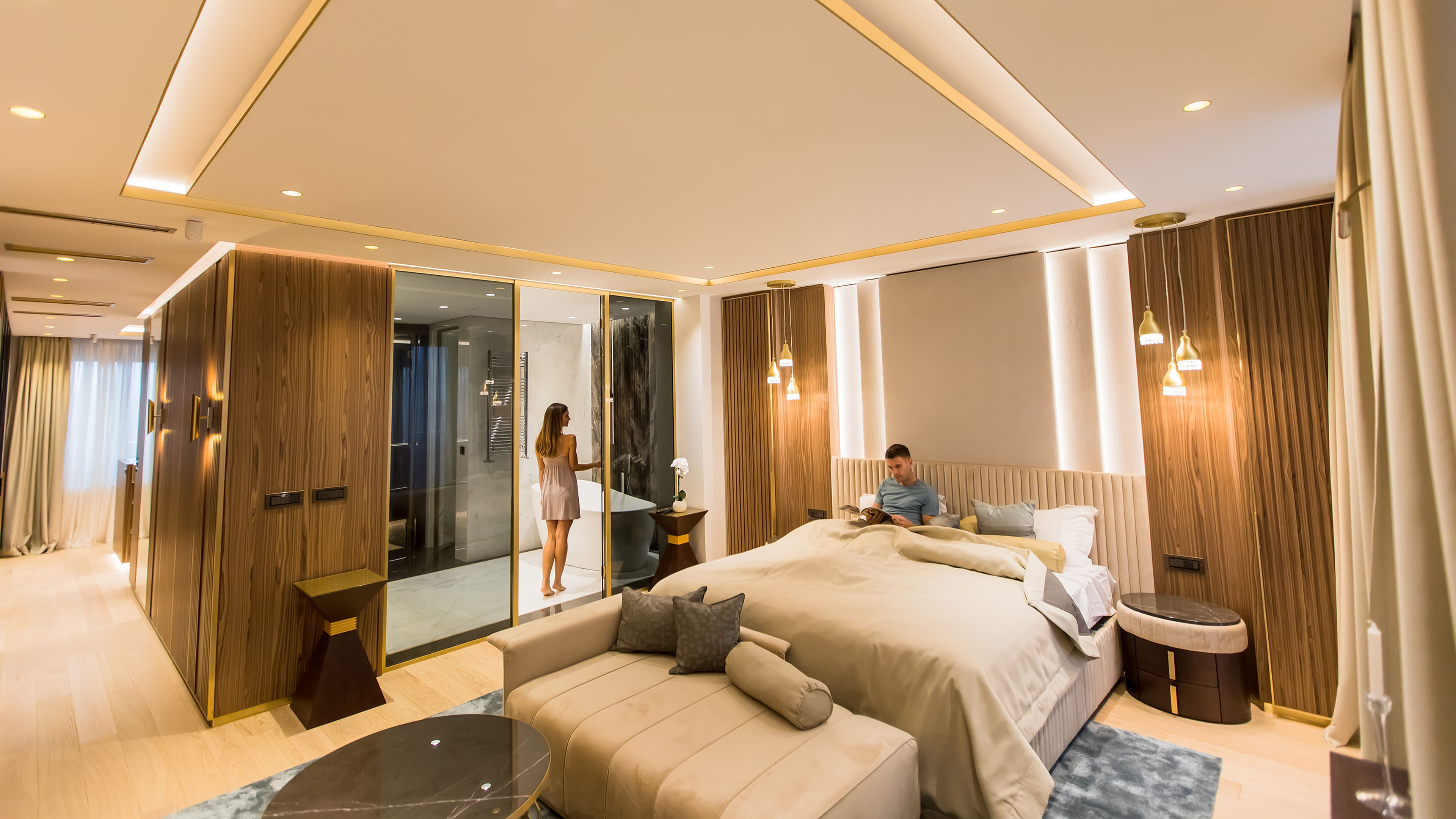 Young couple in a luxurious apartment, with the man lying on the bed and the woman standing inside the room.