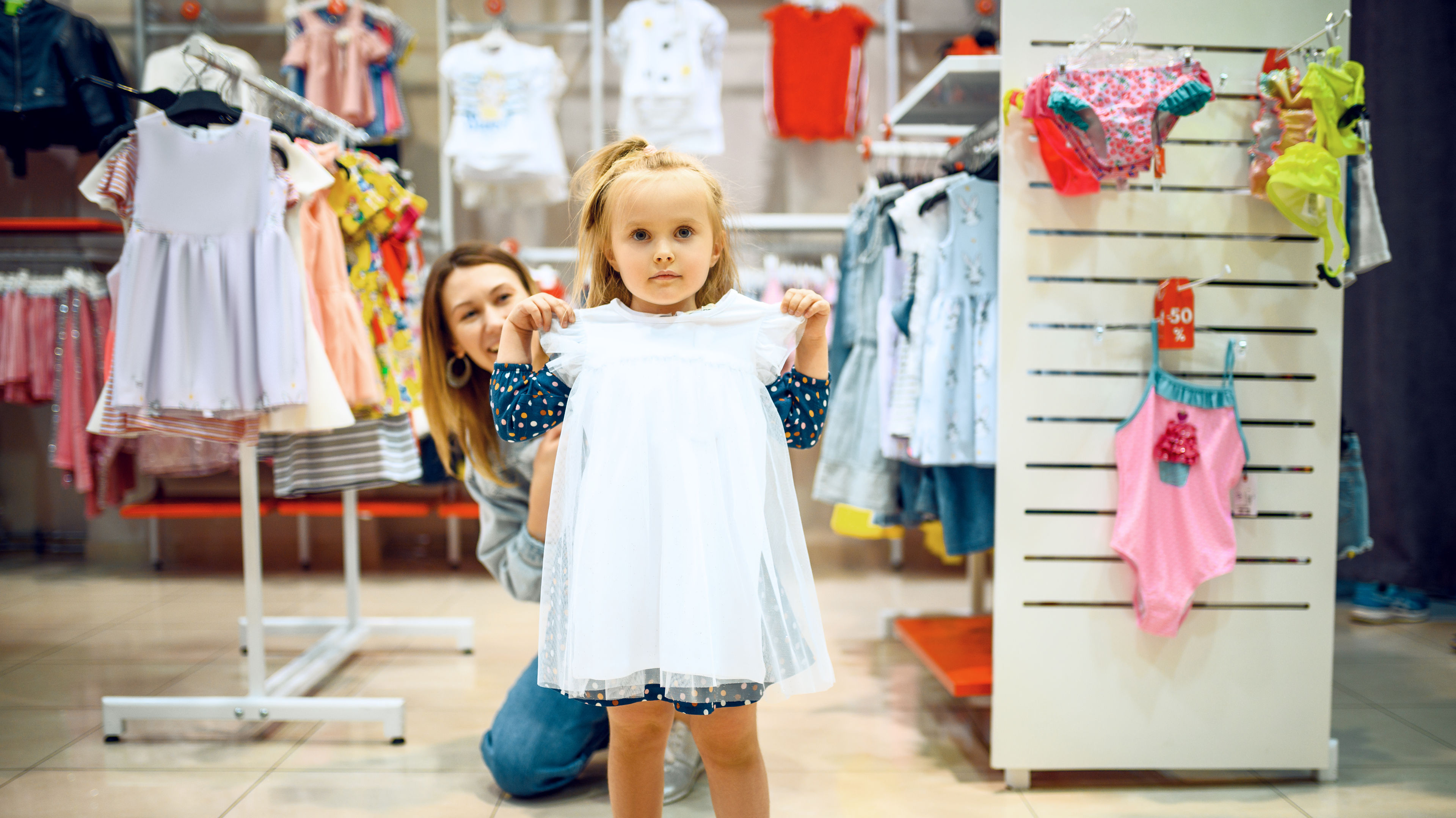 A little girl holding up a white dress in a clothing store, with her mother smiling in the background.