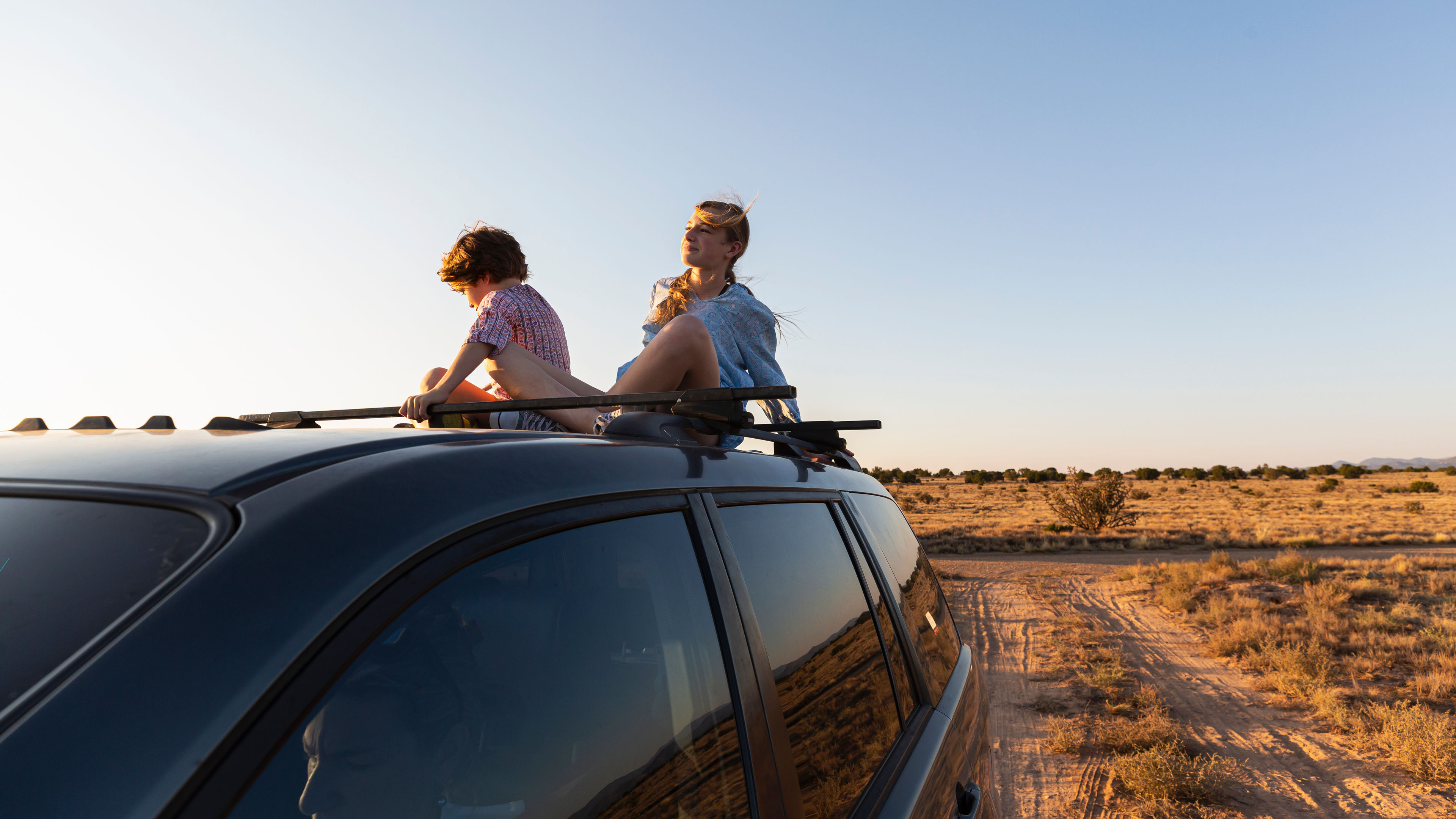 A teenage girl and her younger brother sitting on the roof of a vehicle on a desert road.