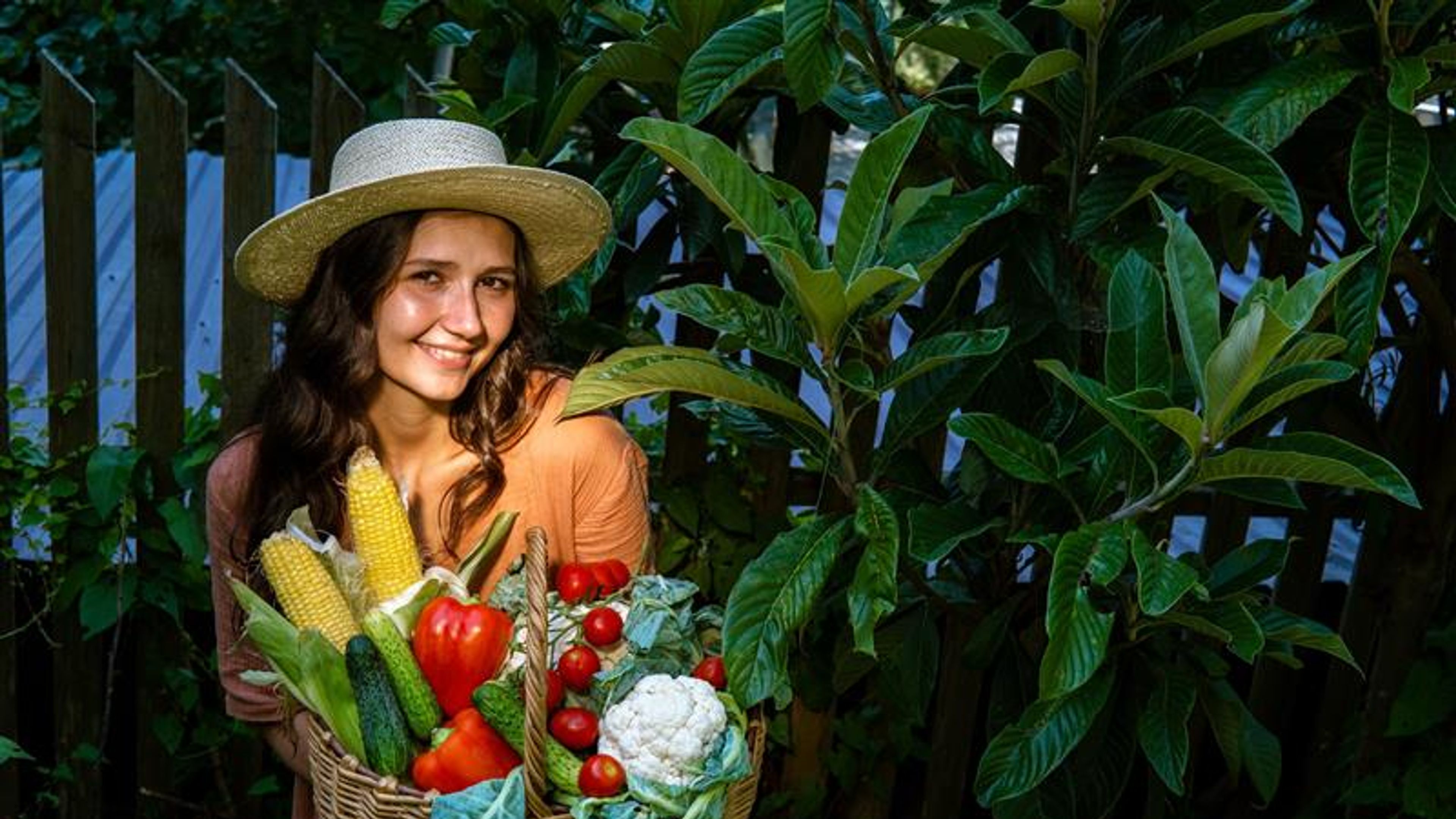 Beautiful woman wearing straw hat and linen dress in garden with basket full of vegetables